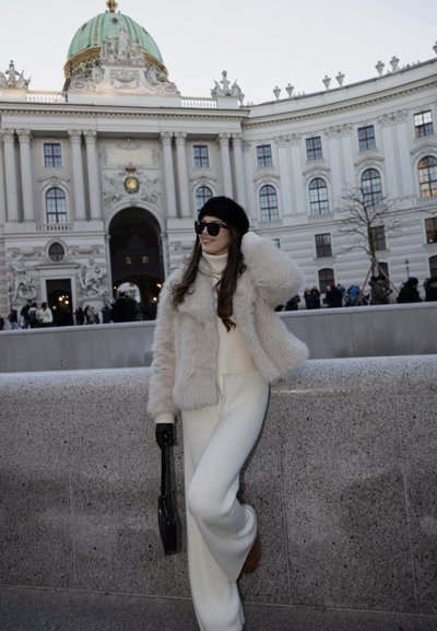 Chaqueta de piel sintética en color crema, suéter blanco con textura acanalada y pantalones de corte ancho. Guantes y bolso negros. De fondo, un edificio ornamentado con una cúpula verde.
