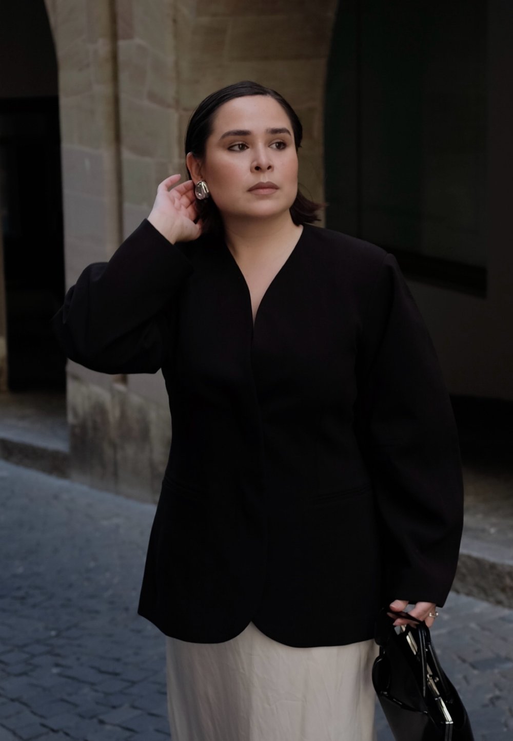 Kobieta w czarnym blezerze i białej spódnicy poprawia kolczyk lewą ręką, w drugiej trzyma czarną torbę, stoi na brukowanej ulicy.