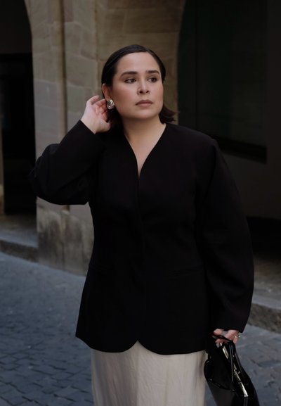 Mujer con blazer negro y falda blanca ajusta un pendiente con la mano izquierda, mientras sostiene un bolso negro con la otra, de pie en una calle empedrada.