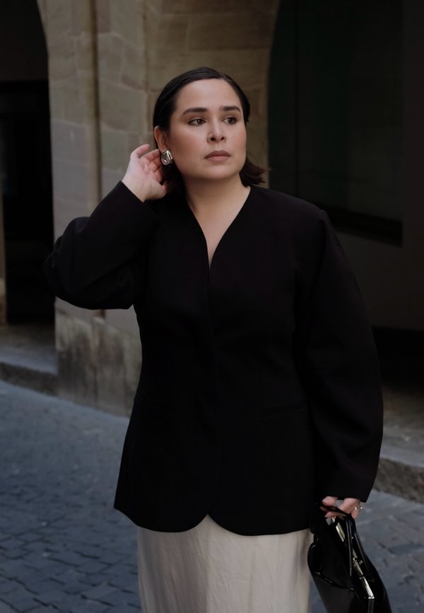 Kvinna i svart kavaj och vit kjol justerar örhänge med vänster hand, håller en svart handväska i den andra, stående på en kullerstensgata.