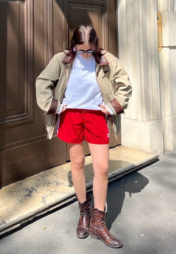 Groene jas met bruine accenten, witte t-shirt, rode shorts en bruine enkellaarsjes. Staande op een stenen oppervlak nabij bruine deuren.