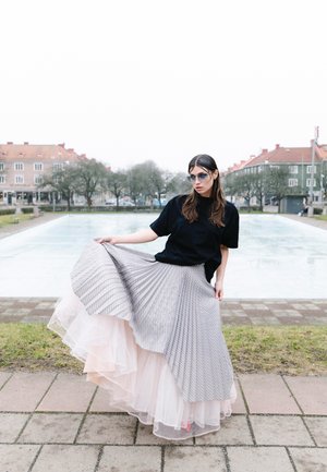 Femme portant des lunettes de soleil, un t-shirt noir et une jupe plissée en couches se tient près d'un étang dans un parc urbain avec des bâtiments et des arbres en arrière-plan.