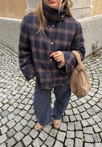 Veste à carreaux bleu marine avec col montant, sous-pull beige à col roulé, pantalon foncé et sac tressé beige. Chaussures marron à lacets. Fond pavé.