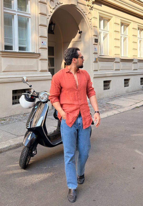 Hombre con una camisa naranja y vaqueros azules camina por una calle junto a un scooter negro con un casco blanco colgado del manillar.