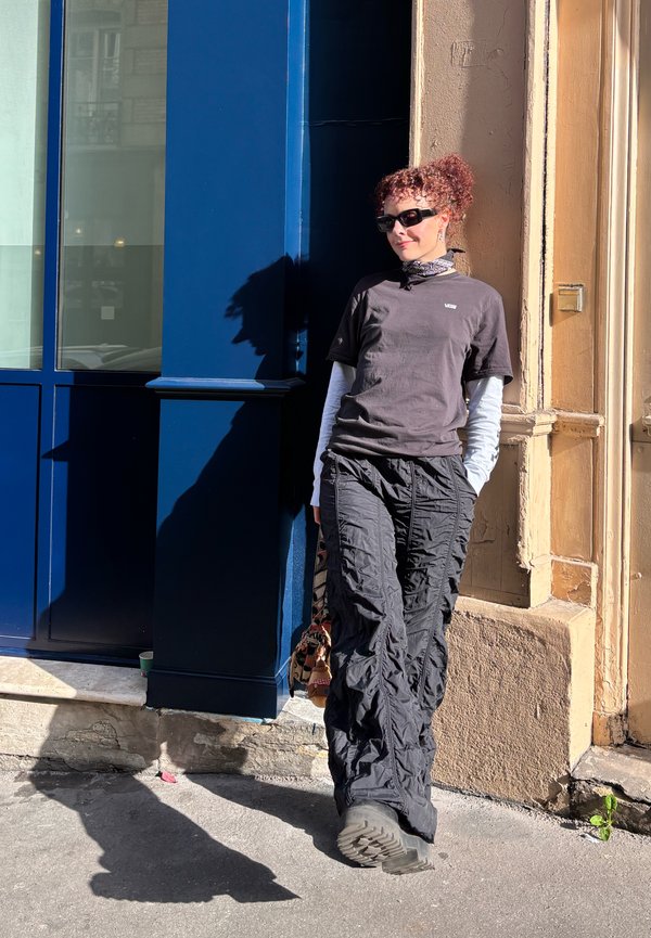 Persona con cabello rizado, gafas de sol, camiseta negra de Vans, mangas largas en capas y pantalones cargo negros apoyada contra un edificio beige y azul bajo la luz del sol.