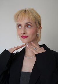 Collier en maille argentée, boucles d'oreilles en argent en spirale, bagues avec des pierres précieuses, veste noire. Le mannequin a les cheveux blonds clairs et porte une couleur de lèvres rose pâle.