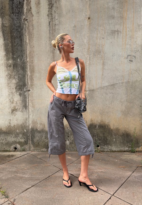 Femme portant un débardeur orné de dentelle, un pantalon gris à jambes larges, des sandales à talons, des lunettes de soleil et portant un petit sac à main noir, debout contre un mur en béton usé.