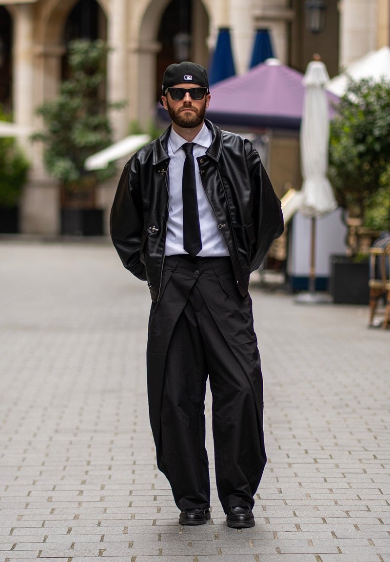 Homme portant une veste en cuir noire, une chemise blanche, une cravate noire, un pantalon large noir, une casquette noire et des lunettes de soleil marchant sur une rue pavée.