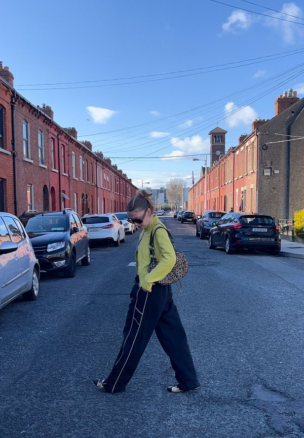 Žena v slnečných okuliaroch, žltej bunde a čiernych nohaviciach kráčajúca cez ulicu lemovanú zaparkovanými autami a radovými domami z červených tehál pod modrou oblohou.