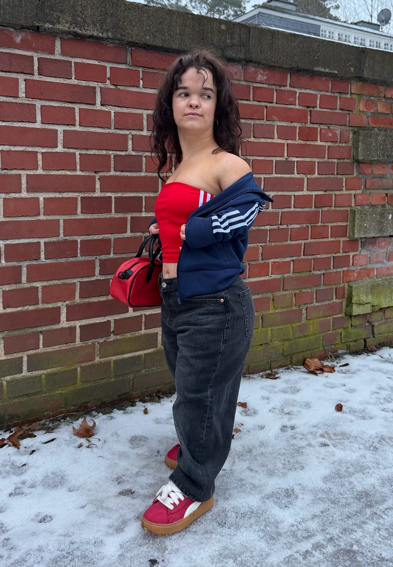 Jeune femme portant un top bandeau rouge, une veste bleue, un jean noir et des baskets rouges, debout sur un sol glacé près d'un mur en briques, tenant un sac à main rouge.
