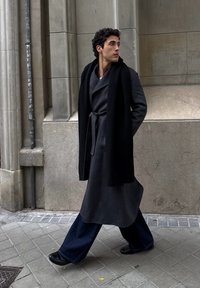 Jeune homme marchant sur le trottoir vêtu d'un long manteau gris foncé ceinturé, d'une écharpe noire, d'un pantalon large bleu marine et de chaussures noires, avec un mur en pierre en arrière-plan.