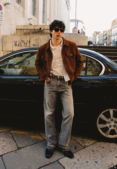 Chaqueta de ante marrón sobre una camisa blanca, jeans azul desgastados y zapatos negros. De pie junto a un coche negro en un entorno urbano.