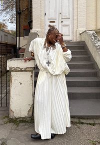 Hvid plisseret kjole med indsnøring i taljen, lange ærmer og løs pasform. Tilbehør i form af smykker og sorte sko. Står på stentrapper.