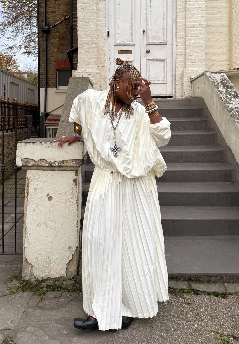 Hvit plissiert kjole med innsvinget midje, lange ermer og løs passform. Tilbehør med smykker og svarte sko. Står på steintrapper.