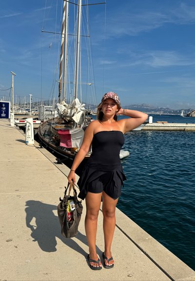 Vestido negro sin mangas con falda acampanada, usado con chanclas. Una gorra de leopardo, que sostiene un bolso oscuro, está de pie en un muelle junto al agua.