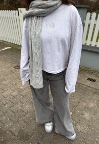 Pelēks īsais džemperis ar izšūtu logotipu, saskaņots ar plašām pelēkām džinsa biksēm un adītu pelēku kabelveidīgu šalli. Sudrabainas slip-on kurpes.