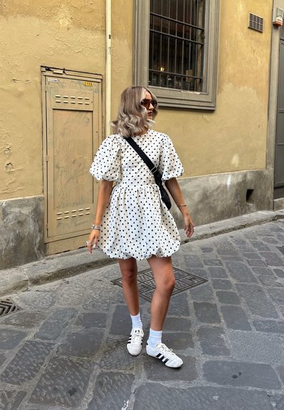 Vestido blanco con lunares con mangas abullonadas y falda fruncida. Lleva un bolso cruzado negro, calcetines blancos y zapatillas blancas con rayas negras.