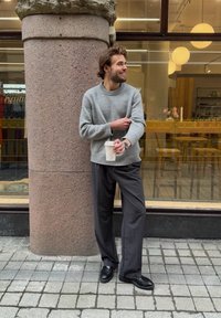 Suéter cinza de malha, calças pretas às riscas, sapatos pretos; segurando uma chávena de café, de pé contra um pilar de pedra, com um fundo de café.