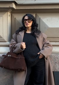 Geantă de mână din piele întoarsă maro cu detaliu cu cataramă, pulover negru supradimensionat și o haină bej. Sunt vizibile și părul lung, ondulat și ochelari de soare mari.