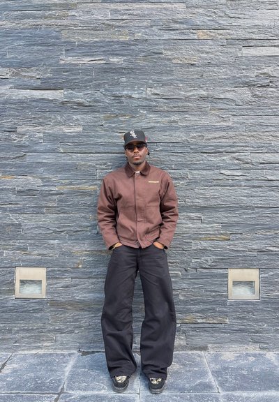 Hombre con chaqueta marrón, pantalones anchos negros, gorra negra y gafas de sol, de pie contra una pared de piedra gris texturizada con las manos en los bolsillos.