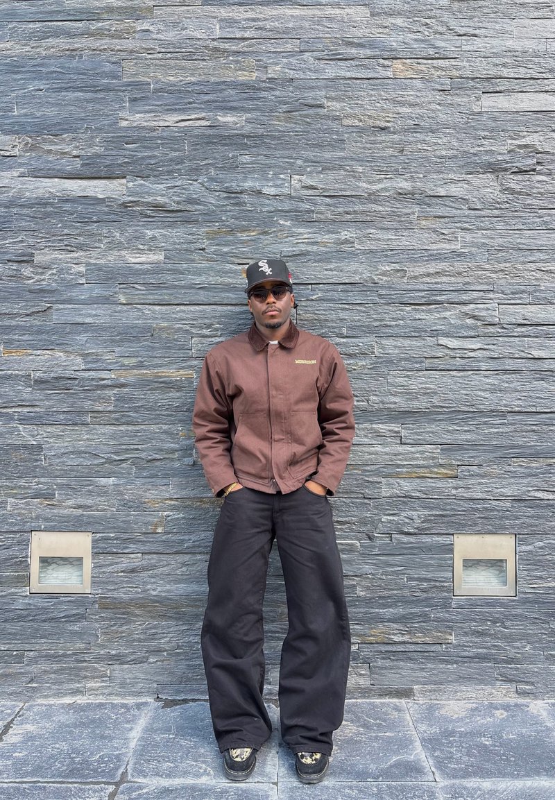 Mees pruunis jopes, mustade laia lõikega pükstega, musta bareti ja päikeseprillidega seisab käed taskutes halli tekstuurse kivi seina ääres.