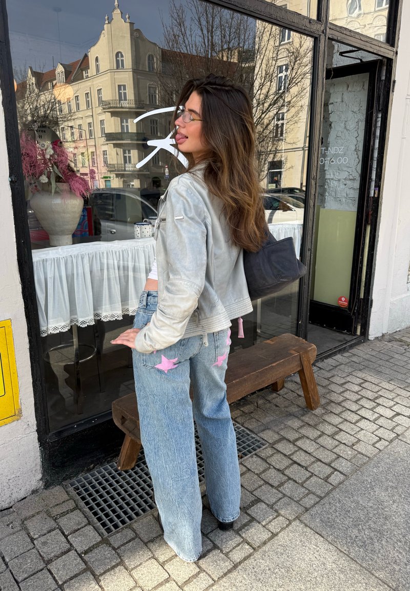 Femme aux cheveux longs et lunettes, portant une veste légère et un jean bleu avec des étoiles roses, debout sur un trottoir près d'une vitrine de café.