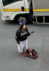 Jeune femme en vêtements décontractés tenant un téléphone, debout à côté d'un grand sac marron sur l'asphalte, près d'un camion blanc avec des rayures jaunes.