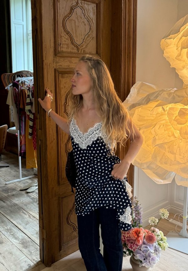 Kvinde med langt blondt hår iført en polkaprikket top og jeans står og holder en trækarm i en stue med tøj og blomster i nærheden.
