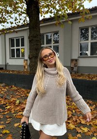 Beige en crèmekleurige gestreepte coltrui met lange mouwen, gedragen over een witte jurk. Zwarte handtas met studs. Achtergrond van herfstbladeren.
