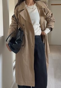 Trench-coat beige à double boutonnage, sac noir à finition brillante, associé à un pull blanc et un pantalon large foncé.