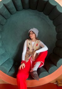 Bežinė liemenė su spalvingomis pleistrais, balta ilgarankovė palaidinė, raudonos kelnės, pilkas beanie, rudi batai, sėdintis apvalioje žalioje pagalvėje.