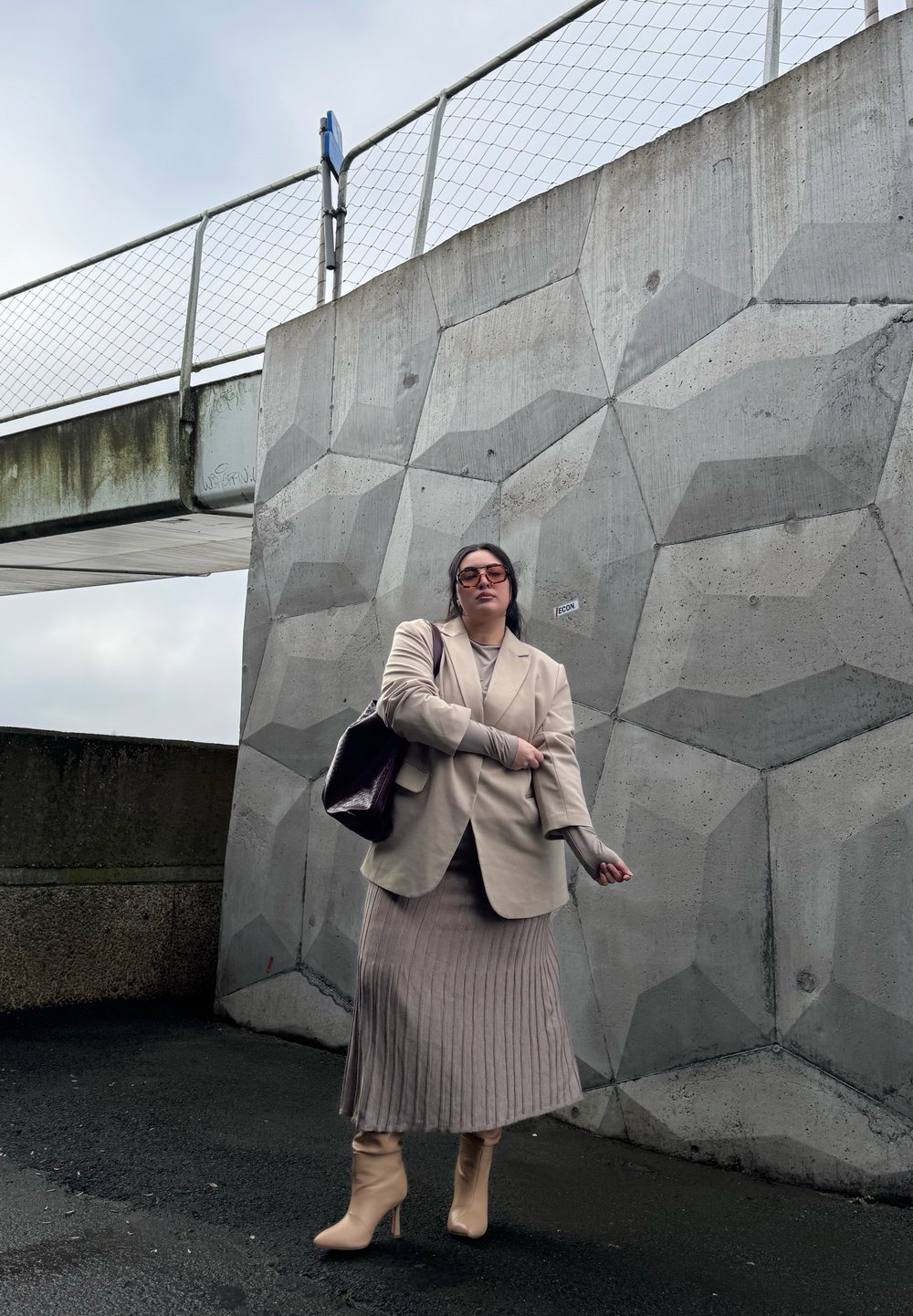Beige skræddersyet blazer over en grå ribbet nederdel, parret med beige hæle og en mørk håndtaske; baggrund med tekstureret betonvæg.