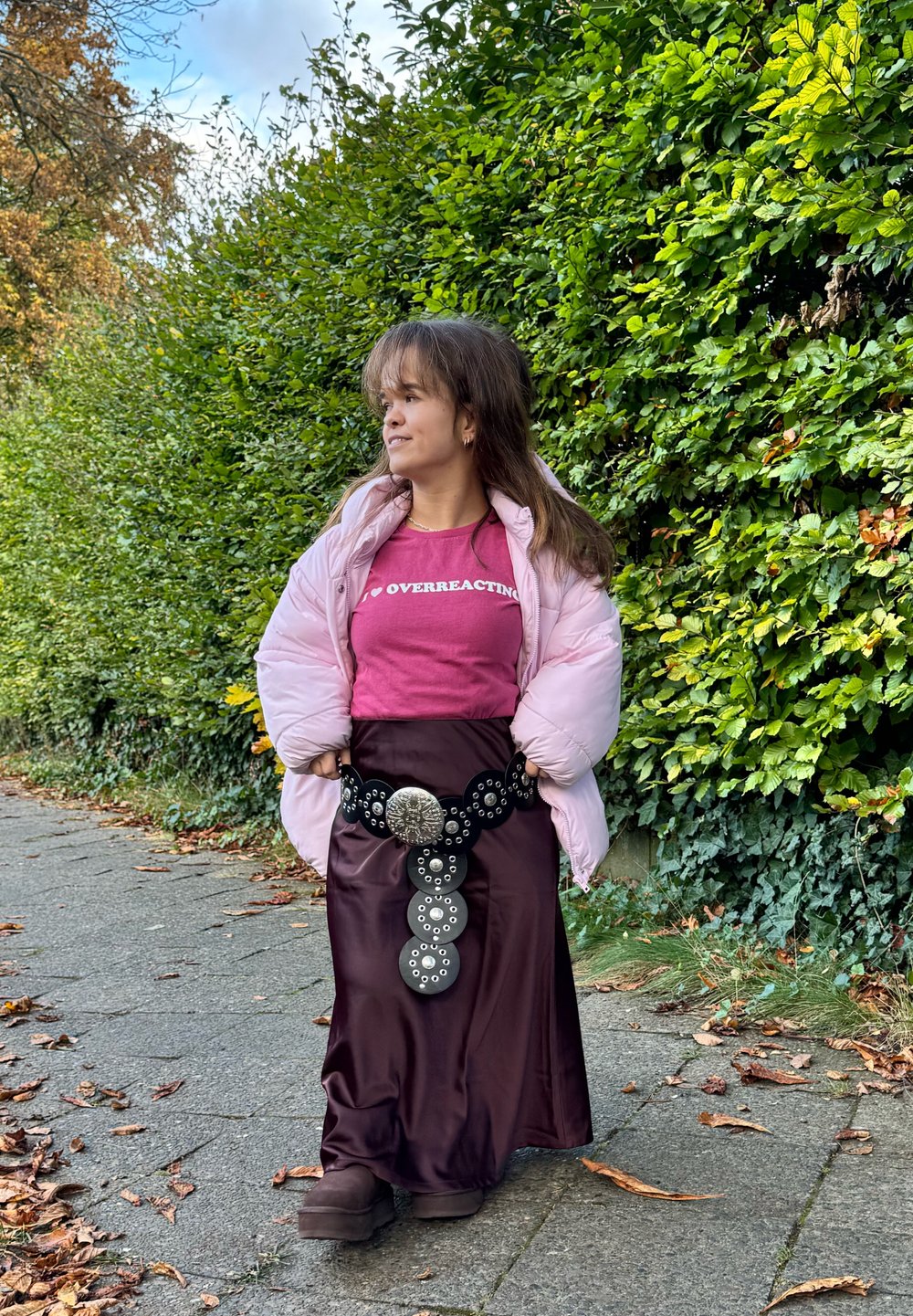 Giacca a vento rosa chiaro sopra una gonna bordeaux, accentuata da una grande cintura nera con dettagli circolari in argento. Stivali marroni. Sfondo di siepe verde.