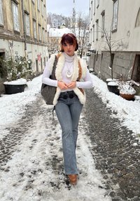 Personne portant un col roulé blanc, un gilet marron et crème, et un jean évasé à rayures fines. Les accessoires comprennent un collier en spirale et un sac gris.