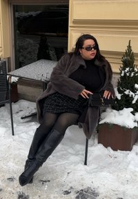 Γυναίκα με σκούρα γυαλιά ηλίου, παλτό από γούνα και μπότες, καθισμένη σε καρέκλα σε εξωτερικό χώρο σε χιονισμένο αστικό περιβάλλον, κρατώντας μια μαύρη τσάντα κοντά σε ένα φυτό καλυμμένο με χιόνι.