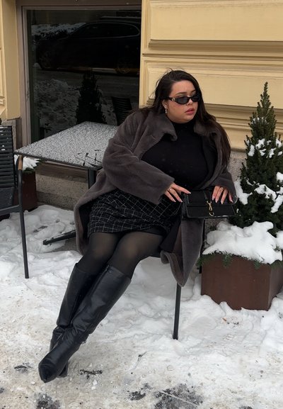 Mujer con gafas de sol oscuras, abrigo de piel y botas sentada en una silla al aire libre en un entorno urbano nevado, sujetando un bolso negro cerca de una planta cubierta de nieve.