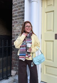 Femme avec des lunettes portant un pull jaune et une écharpe à motifs, tenant une tasse de café et un sac à main bleu clair, debout près d'un mur en brique et d'une porte jaune pâle.