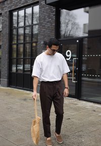 Vit kortärmad skjorta, mörkbruna avsmalnande byxor, bruna slip-on skor och en vävd beige väska, mot en tegelvägg och glasdörr.