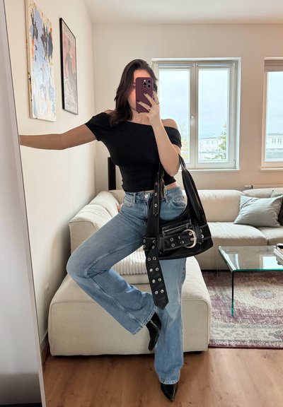 Blusa negra sin mangas, vaqueros acampanados de color azul claro, bolso negro con herrajes metálicos, de pie frente a un espejo con un sofá detrás.