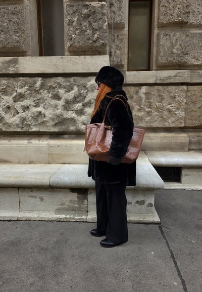 Mujer con abrigo de piel negro, sombrero y guantes, llevando un gran bolso marrón, de pie en la acera junto a un edificio de piedra texturizada.