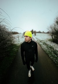 Coureur en tenue noire et chapeau jaune vif debout sur un sentier enneigé, regardant vers la gauche, entouré d'arbres nus et d'un paysage hivernal.