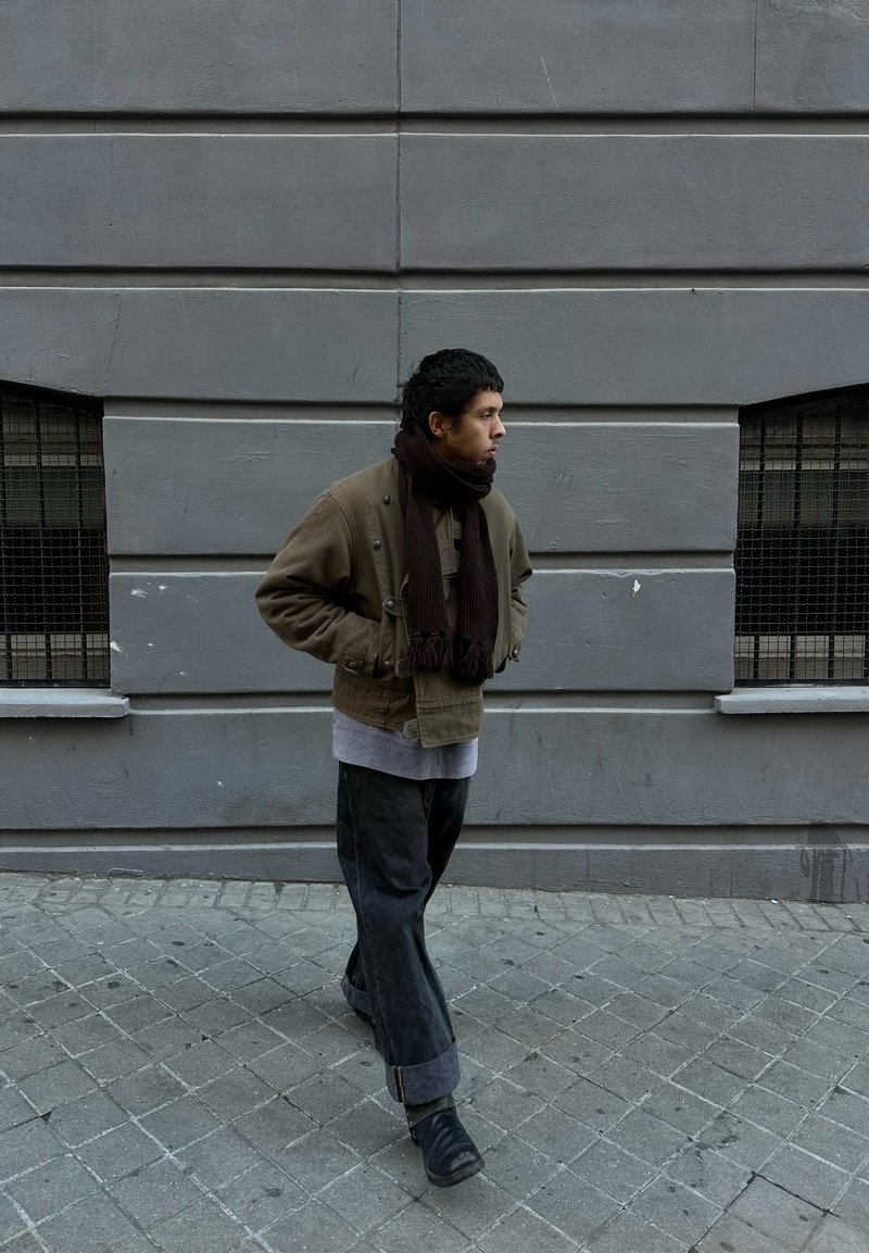 Brown military-style jacket with buttons, dark denim trousers, and a dark scarf. Walking on grey pavement, with a textured grey wall in the background.