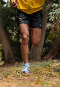 Czarne spodenki do biegania w wzory z pomarańczowymi akcentami, zestawione z jasnoniebieskimi butami do biegania. W tle widać zieleń i miękką nawierzchnię.