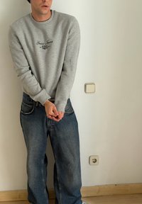 Muž v šedé mikině Guess Jeans a volných modrých džínách stojí uvnitř, ruce sepnuté před jednobarevnou zdí s elektrickými zásuvkami.