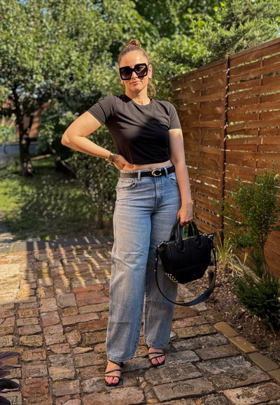 Camiseta corta negra, jeans de pierna ancha azul claro, bolso negro con tachuelas, gafas de sol de gran tamaño y sandalias negras sobre un sendero de piedra.