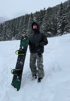 Persona con chaqueta negra, pantalones estampados y gafas de color rosa sosteniendo una tabla de snowboard verde y negra, de pie sobre la nieve cerca de pinos.