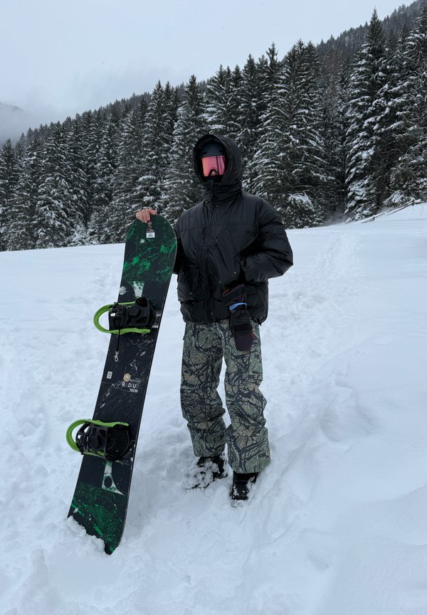 Persona con giacca nera, pantaloni a fantasia e occhiali rosa che tiene una tavola da snowboard verde e nera, in piedi sulla neve vicino agli alberi di pino.