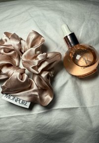 Satiininen scrunchie pehmeässä vaaleanpunaisessa värissä tarran kera, vieressä pyöreä lasipullo hiöljyä tiputuspipetillä, jossa on kultainen yksityiskohta ja logo.