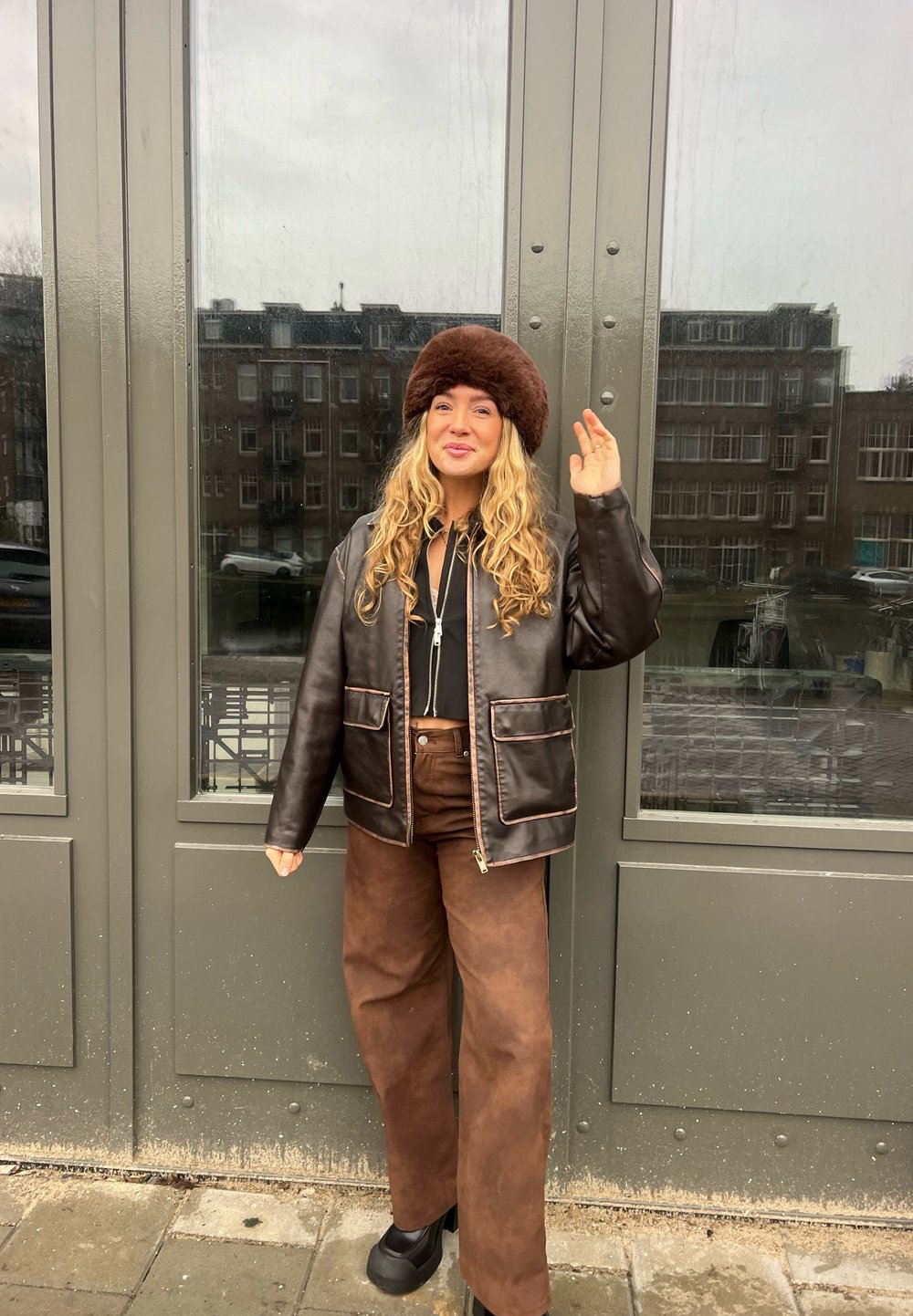 Giubbotto in pelle marrone con tasche frontali e cuciture a contrasto, abbinato a pantaloni larghi marroni e a un cappello di pelliccia marrone.