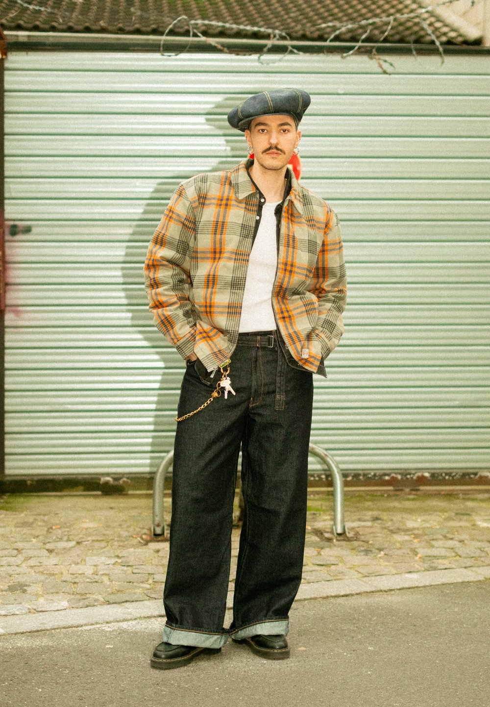 Veste à carreaux vert, orange et beige, associée à un pantalon large en denim foncé. Chaîne d'accent avec un pendentif, et un béret bleu.